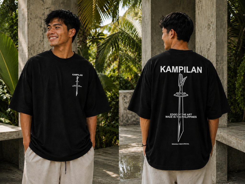 A man wearing a black oversized T-shirt shown from both front and back in an outdoor patio setting with stone pillars and greenery. The front has a small white sword graphic and text, while the back has a large white sword graphic and the word 'KAMPILAN'.